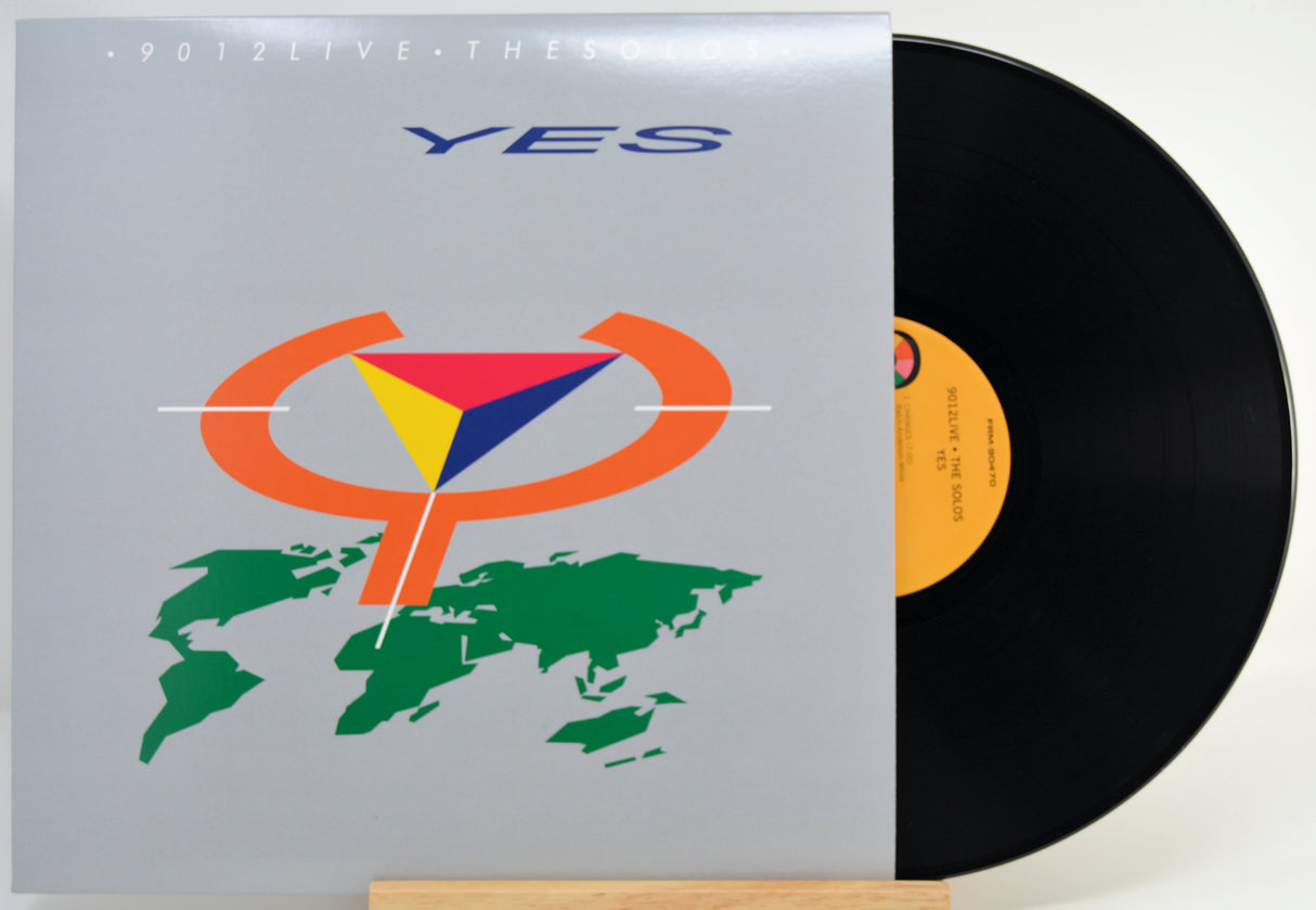 A vinyl record partially pulled from its sleeve, showing the front cover artwork for Yes's '9012 Live' album. The sleeve is light gray with blue 'YES' text and a multicolor geometric logo over a green world map.