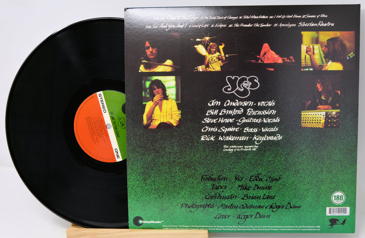 A vinyl record partially pulled from its sleeve, showing the black record on the left and the back cover of the album on the right. The sleeve features a collage of photos and text, with a green-to-black gradient background.