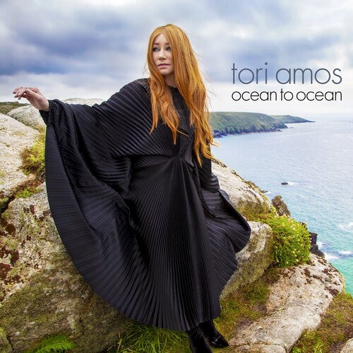 Tori Amos sitting on a rocky outcrop by the sea, wearing a black, flowing outfit.