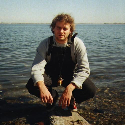 A person crouching by a body of water, wearing a white shirt and black overall, with a watch and a lanyard around the neck.