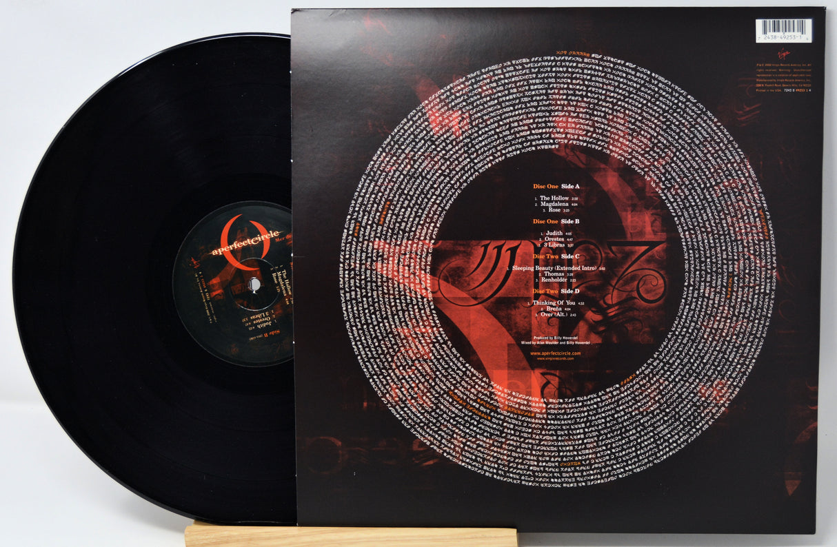 A black vinyl record partially pulled from its sleeve, displayed on a small wooden stand against a light gray background. The album cover is predominantly black with a large circular white text graphic and red abstract artwork.
