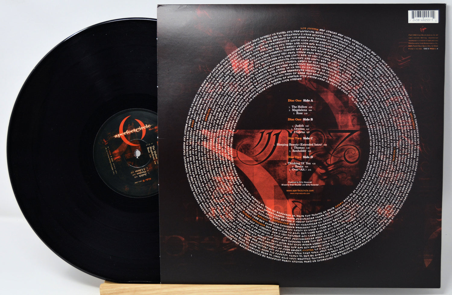A black vinyl record partially pulled from its sleeve, displayed on a small wooden stand against a light gray background. The album cover is predominantly black with a large circular white text graphic and red abstract artwork.
