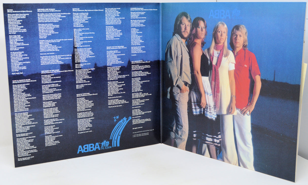 An open ABBA 'The Single' vinyl record album, showing the printed tracklist on the left panel and the band members standing against a blue sky background on the right panel.