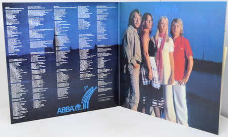 An open ABBA 'The Single' vinyl record album, showing the printed tracklist on the left panel and the band members standing against a blue sky background on the right panel.