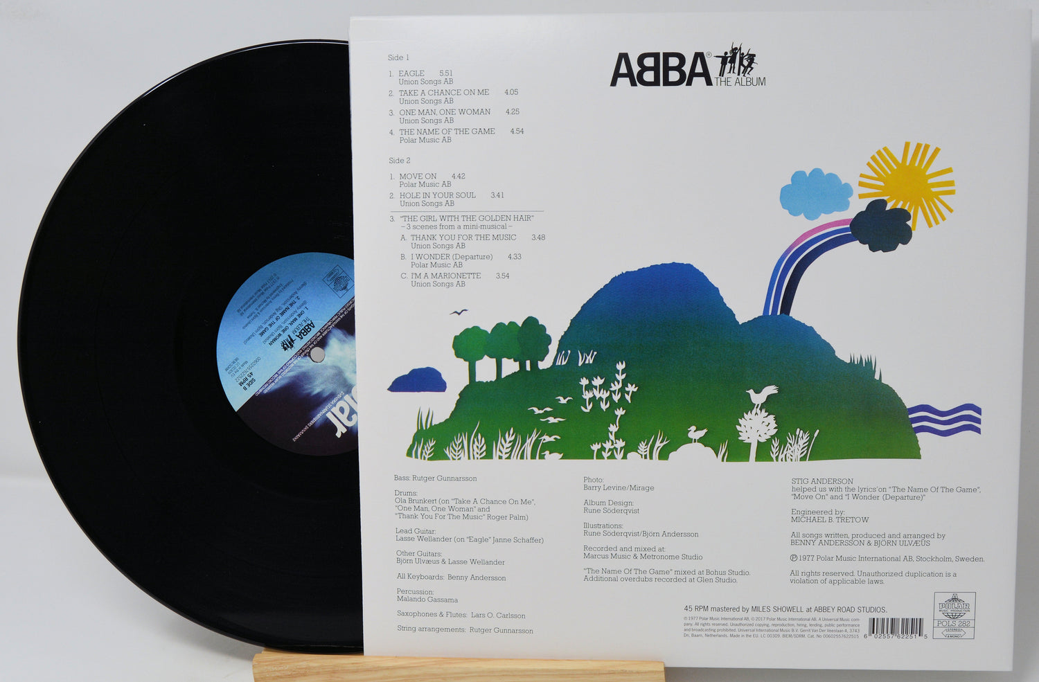 ABBA The Album vinyl record shown with the black LP partially pulled from the printed back cover, which features a colorful landscape illustration and track listing.
