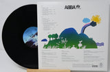 ABBA The Album vinyl record shown with the black LP partially pulled from the printed back cover, which features a colorful landscape illustration and track listing.