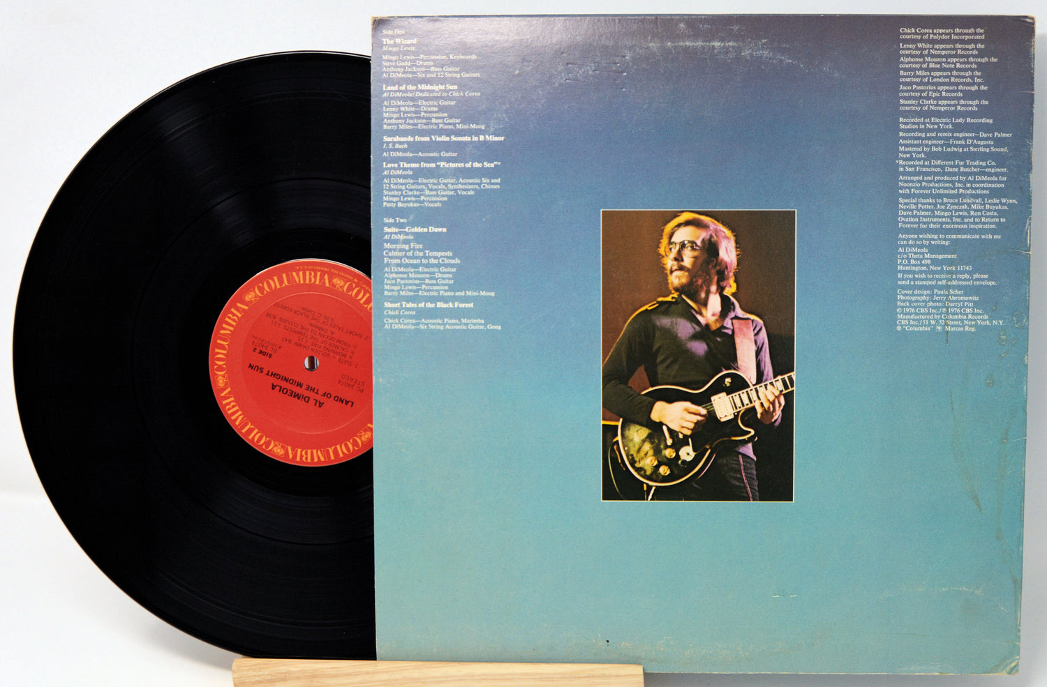 A vinyl record partially pulled from its sleeve, showing the black record with a red center label and the back cover featuring a blue gradient and a small photo of a musician playing an electric guitar.