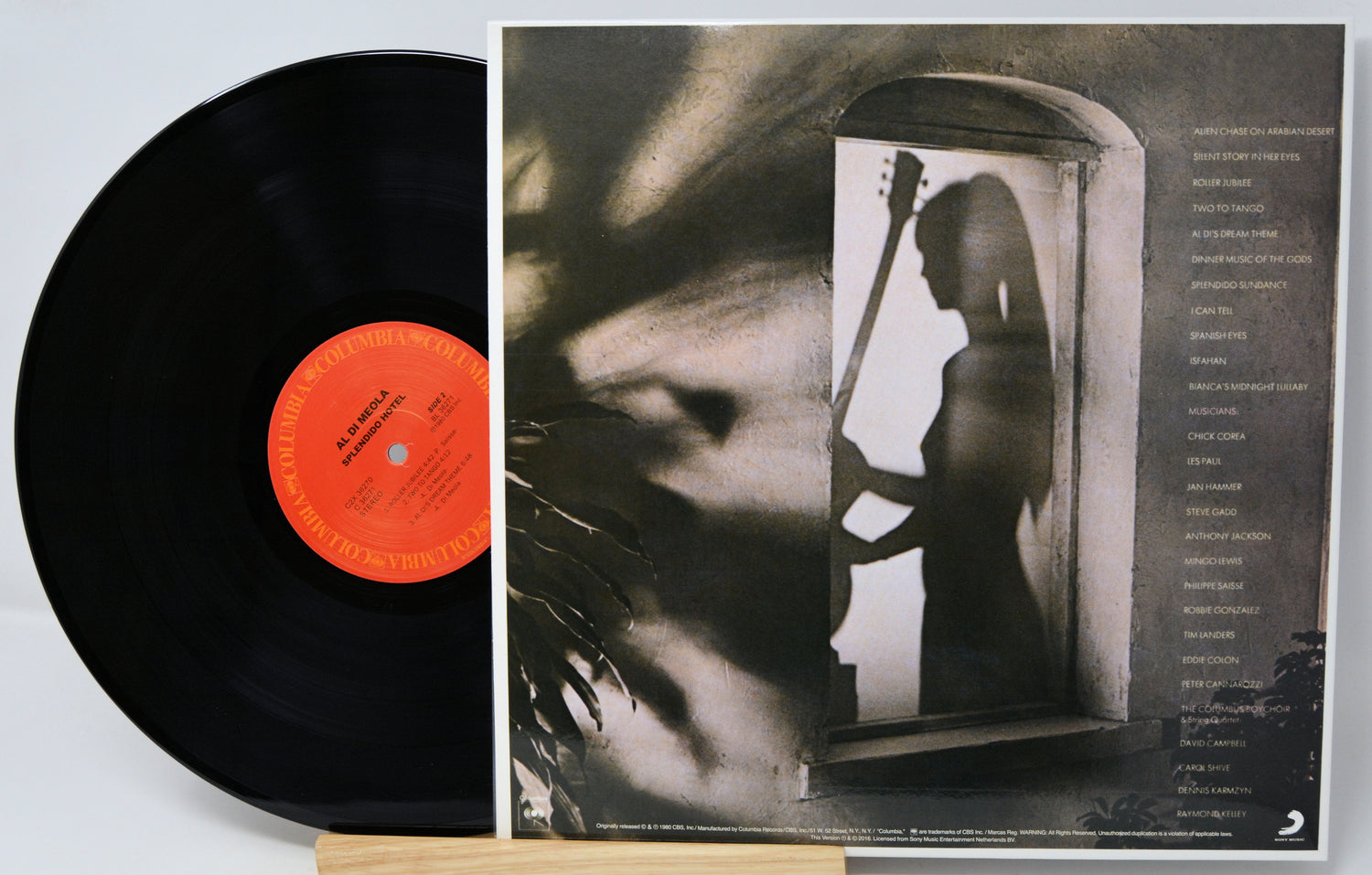 A vinyl record partially pulled from its sleeve, showing the black record with a bright orange center label and the printed album cover behind it. The cover features a monochrome photo of a woman holding a stringed instrument in a doorway, with tracklist text along the right side.