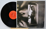 A vinyl record partially pulled from its sleeve, showing the black record with a bright orange center label and the printed album cover behind it. The cover features a monochrome photo of a woman holding a stringed instrument in a doorway, with tracklist text along the right side.