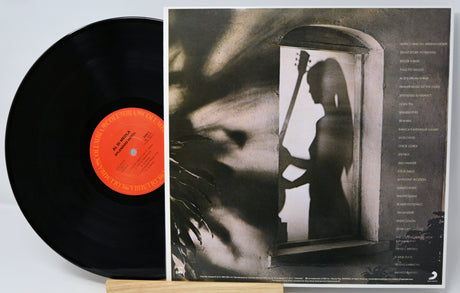 A vinyl record partially pulled from its sleeve, showing the black record with a bright orange center label and the printed album cover behind it. The cover features a monochrome photo of a woman holding a stringed instrument in a doorway, with tracklist text along the right side.