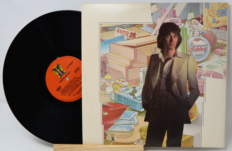 A vinyl record partially pulled from its sleeve, showing the black record with an orange center label and the illustrated album cover featuring a man in a beige suit against a collage-style background.