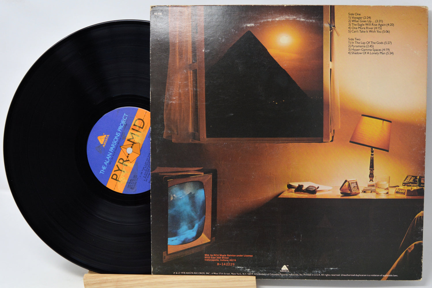 A vinyl record partially pulled from its album cover, displayed on a small wooden stand against a white background. The cover art depicts a warm-toned room with a television and a lamp, and the record label features a blue and orange pyramid graphic.