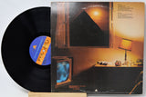A vinyl record partially pulled from its album cover, displayed on a small wooden stand against a white background. The cover art depicts a warm-toned room with a television and a lamp, and the record label features a blue and orange pyramid graphic.