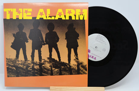 A vinyl record titled 'The Alarm' with a dark cover featuring silhouettes of people. The record is placed on a wooden platter.