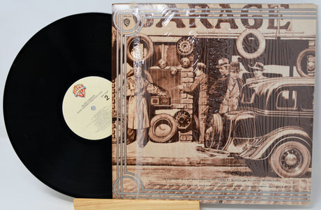 Vinyl record with a detailed cover featuring vintage cars and people, on a light gray background.