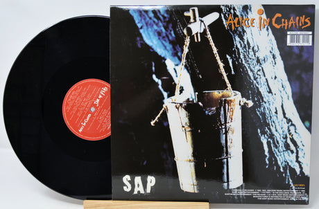 A vinyl record and its album cover for Alice in Chains's 'SAP' on a light background. The black record is partially pulled from the cover, which features the band's name in orange at the top right and the album title 'SAP' in large white letters at the bottom left.