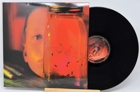 Vinyl record album with the cover partially pulled back to reveal a black LP disc. The cover art shows a close-up of a face behind a large glass jar filled with orange-red liquid and small insects, photographed against a light gray background.