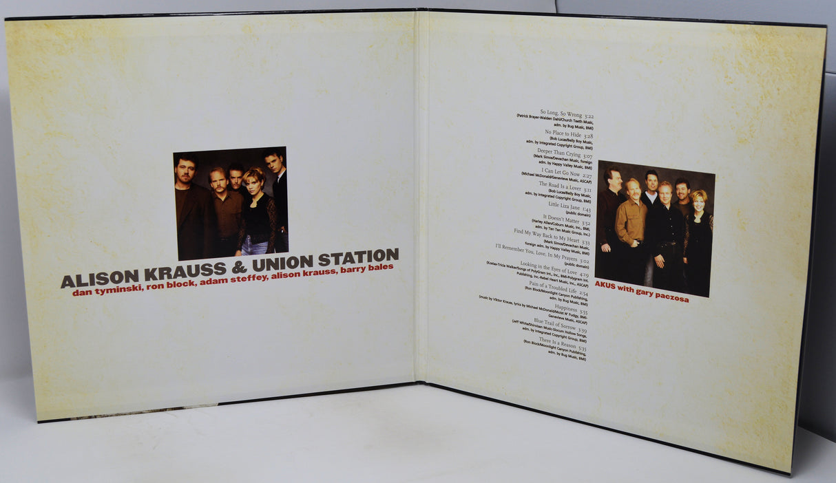 An open printed booklet or album insert with two cream-colored pages, each featuring a small black-and-white band photo and text. The left page reads 'ALISON KRAUSS & UNION STATION' with smaller text below, and the right page contains song text and a small band photo.