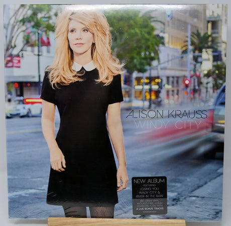 Alison Krauss 'Windy City' album cover, featuring a blonde woman in a black dress standing on a city street. The cover includes a black 'New Album' sticker and the artist's name and album title in white text.