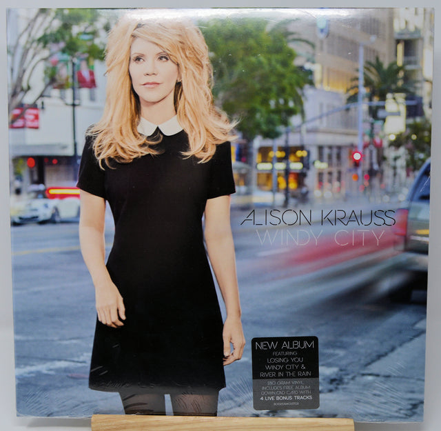 Alison Krauss 'Windy City' album cover, featuring a blonde woman in a black dress standing on a city street. The cover includes a black 'New Album' sticker and the artist's name and album title in white text.