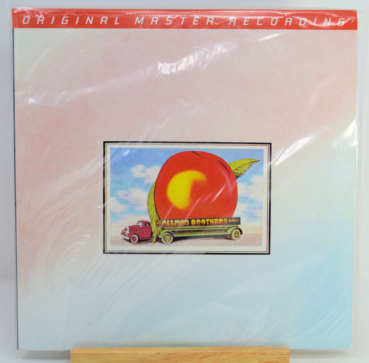The album cover of 'Original Master Recording' by the Allman Brothers, featuring a red and light blue gradient background with a central illustration of a red apple on a green truck. The album is sealed in plastic shrink wrap and displayed on a wooden stand.