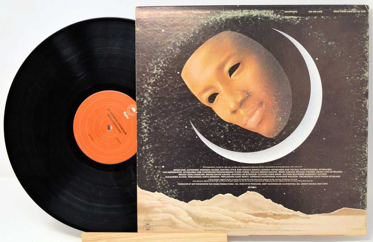 A vinyl record partially pulled from its album cover, showing a black record with an orange center label beside the cover art featuring a stylized face inside a crescent moon.