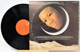 A vinyl record partially pulled from its album cover, showing a black record with an orange center label beside the cover art featuring a stylized face inside a crescent moon.