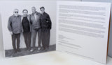 An open printed booklet or book spread showing a black-and-white group photograph of four men on the left page and columns of small text on the right page, photographed against a light neutral background.