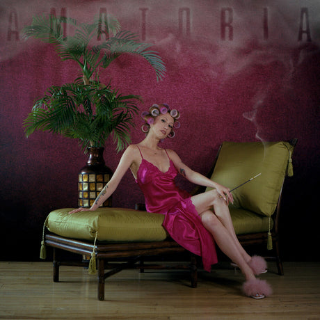 Woman in a pink dress with curlers sitting on a green couch against a dark wall with 'AMATORIA' text.