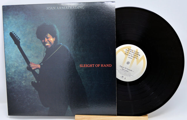 A vinyl record titled 'Sleight Of Hand' by Joan Armatrading with the album cover showing the artist's face and a guitar. The record is placed in front of the cover.