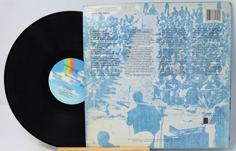 A vinyl record partially pulled from its back cover sleeve, photographed on a plain light background. The record is black with a blue-and-white center label featuring a rainbow arc.