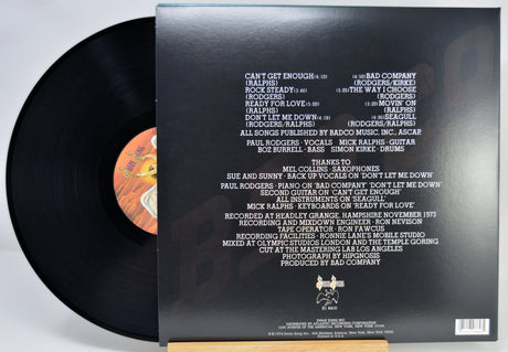 A vinyl record partially pulled from its black outer sleeve, showing the back of the packaging with track listings and a small orange label visible on the black record.