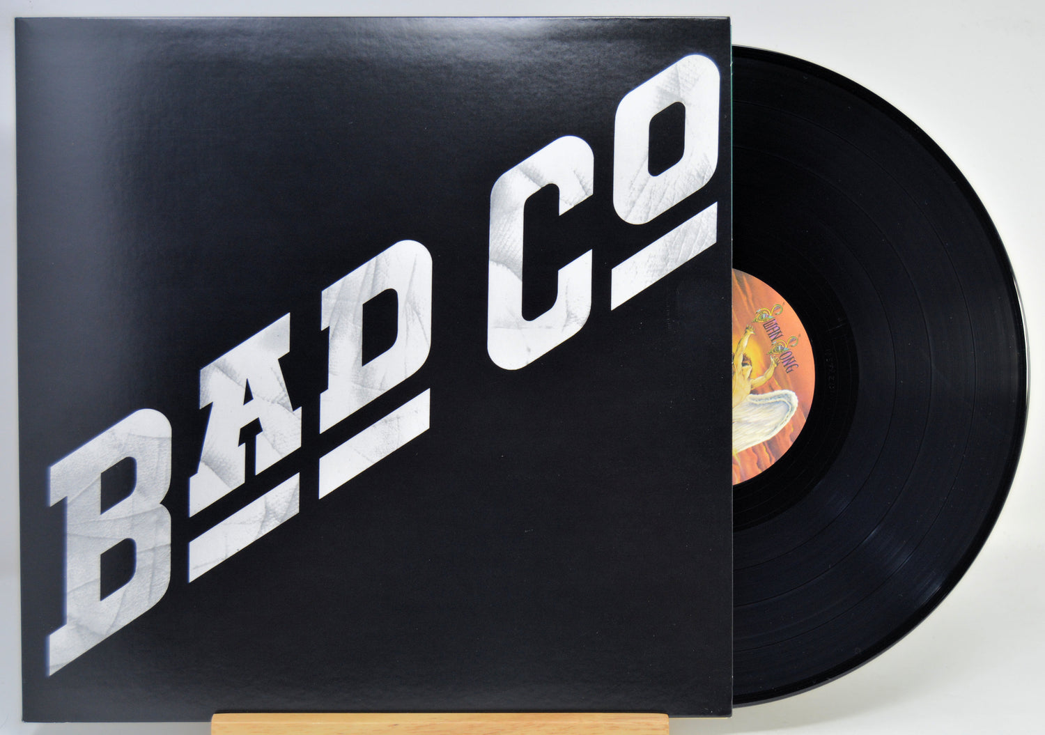 A vinyl record partially pulled from its sleeve, showing the black record and a black cover with large white 'BAD CO' lettering. The center label is visible with a yellow/orange background.