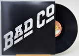 A vinyl record partially pulled from its sleeve, showing the black record and a black cover with large white 'BAD CO' lettering. The center label is visible with a yellow/orange background.