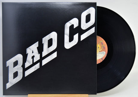 A vinyl record partially pulled from its sleeve, showing the black record and a black cover with large white 'BAD CO' lettering. The center label is visible with a yellow/orange background.