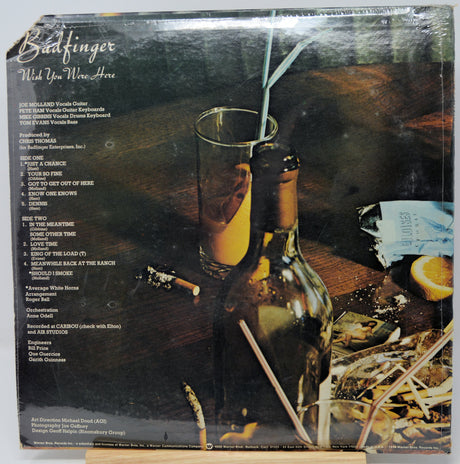 Vinyl record album cover for Badfinger's 'Pick Up Your Wine Here', showing a dark bottle, a glass with an orange drink and straw, a slice of orange, and a messy bar scene. The left side contains the tracklist.