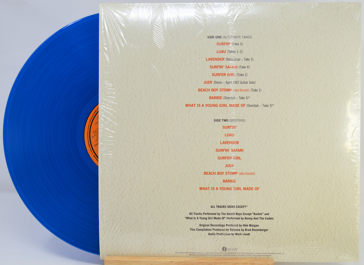 A vinyl record partially pulled from its sleeve, wrapped in clear plastic. The sleeve is cream-colored with orange printed track listings, and the visible record is blue with an orange center label.