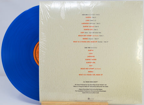 A vinyl record partially pulled from its sleeve, wrapped in clear plastic. The sleeve is cream-colored with orange printed track listings, and the visible record is blue with an orange center label.