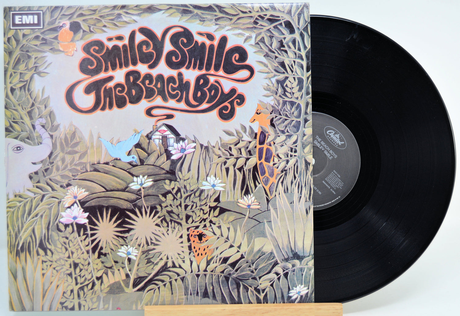 A vinyl record partially pulled from its illustrated album cover, showing the title 'Smiley Smile The Beach Birds' and the EMI logo. The cover features a whimsical jungle scene with animals in muted beige, green, and orange tones, with a black record visible on the right.