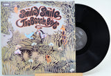 A vinyl record partially pulled from its illustrated album cover, showing the title 'Smiley Smile The Beach Birds' and the EMI logo. The cover features a whimsical jungle scene with animals in muted beige, green, and orange tones, with a black record visible on the right.