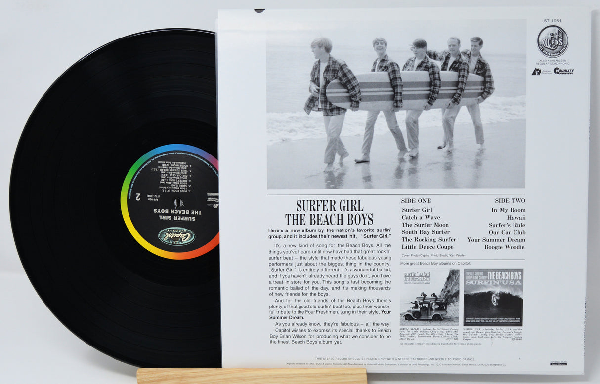 A vinyl record partially pulled from its sleeve, showing the black record with a colorful label and the printed inner sleeve behind it. The sleeve features a black-and-white photo of four surfers walking and track listings for 'Surfer Girl' and 'The Beach Boys'.