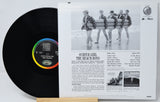 A vinyl record partially pulled from its sleeve, showing the black record with a colorful label and the printed inner sleeve behind it. The sleeve features a black-and-white photo of four surfers walking and track listings for 'Surfer Girl' and 'The Beach Boys'.