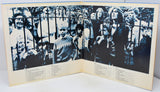 An open vintage book or photo album showing a black-and-white photograph of a crowd behind a fence, with printed text columns on the cream-colored pages.