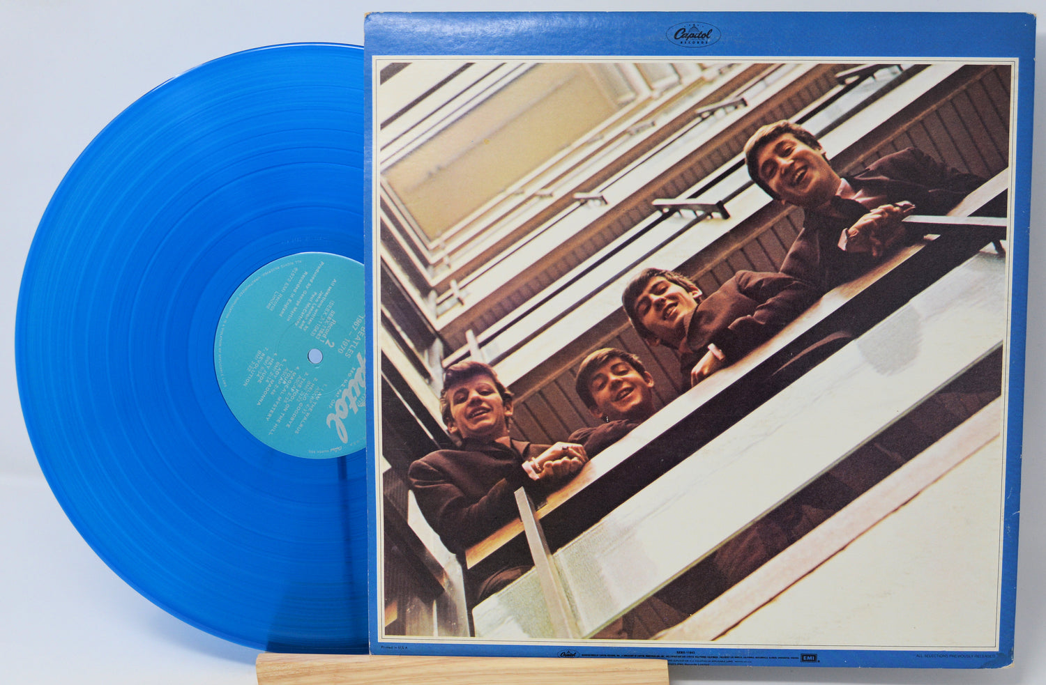 A blue vinyl record partially pulled from its album cover, showing the band members leaning out of a building on the front artwork. The record itself is bright blue with a light blue center label.