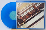 A blue vinyl record partially pulled from its album cover, showing the band members leaning out of a building on the front artwork. The record itself is bright blue with a light blue center label.