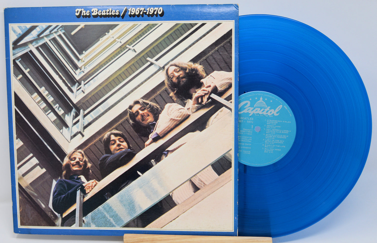 The Beatles '1967-1970' vinyl record album with a translucent blue record partially visible behind the cover, photographed against a light blue background.