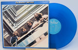 The Beatles '1967-1970' vinyl record album with a translucent blue record partially visible behind the cover, photographed against a light blue background.