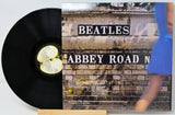 A Beatles 'Abbey Road' vinyl record partially pulled from its album cover, displayed on a small wooden stand against a light gray background.