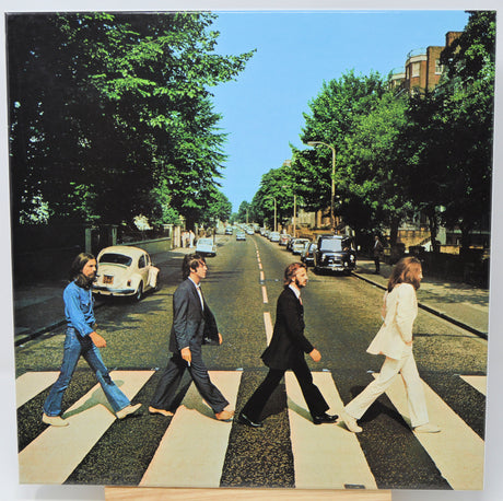 A square art print or poster showing the four Beatles crossing a zebra street in London, with trees, cars, and a clear blue sky in the background. The print is photographed against a white background with a small wooden easel visible at the bottom.