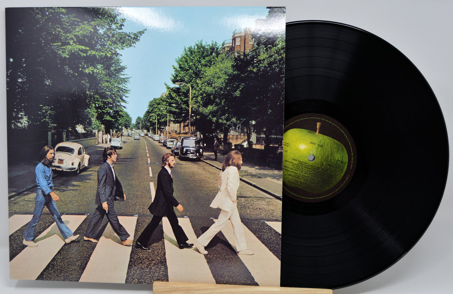 A vinyl record partially pulled from its sleeve, showing the iconic Abbey Road album cover with four men walking across a zebra crossing and a black record disc with a green center label on a light gray background.